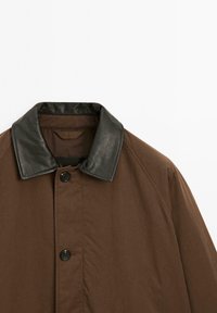 Bruine jas met een zwarte leren kraag, voorzien van knoopsluiting en een gladde, textuurstof. Geen zichtbare patronen of accenten.