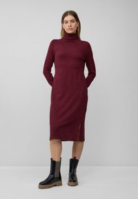Bordeauxfarbendes Rollkragenkleid mit langen Ärmeln, knielang und mit einem seitlichen Schlitz, kombiniert mit schwarzen Ankle-Boots mit strukturierten Seitenpanelen.