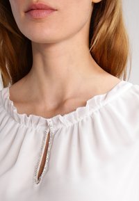 Bílá blůza se skládaným límcem, která má malé otvory ve tvaru klíče a stříbrné lemování podél okraje. Měkká, lehká látka s hladkou texturou.
