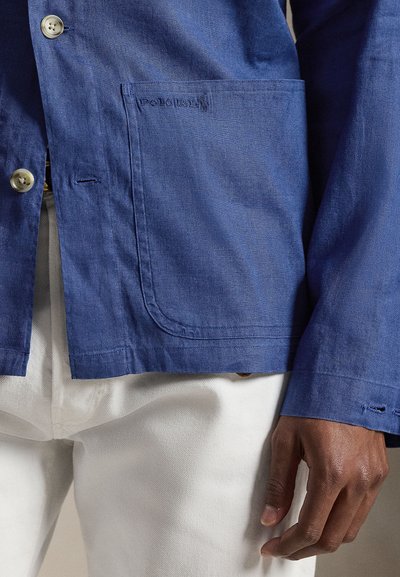 Άνδρας με μπλε σακάκι με κουμπιά και μεγάλη μπροστινή τσέπη, και λευκό παντελόνι, με το χέρι χαλαρό στο πλάι.