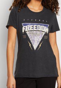 Camiseta de algodón negra con un gráfico triangular que presenta la palabra "LIBERTAD", acentos texturizados y motivos de rayos en blanco y dorado.