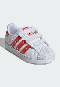 Biele detské tenisky s červenými srdiečkovými akcentmi, tromi pútkami na suchý zips a štruktúrovanou gumovou podrážkou. Obsahujú charakteristický dizajn škrupinovej špičky.