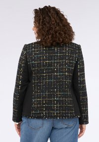 Giacca nera in tweed con degli spruzzi multicolore, spalle strutturate e orlo frangiato. Pannelli laterali in tessuto nero creano un contrasto. Visibili jeans in denim.