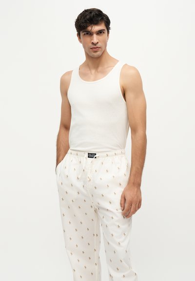 Homme portant un débardeur blanc sans manches et un pantalon blanc avec de petits logos dorés brodés, debout les mains dans les poches.