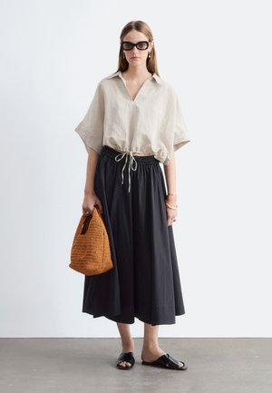 Femme portant une chemise beige oversize, pantalon noir à jambes larges, sandales noires, lunettes de soleil foncées, tenant un sac à main tressé marron sur un sol gris.