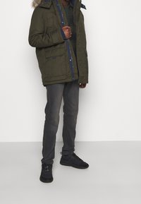 Olivově zelená parka s kapucí zdobenou kožešinou, zapínání na zip a knoflíky, a několika kapsami. Obuté s tmavě šedými džíny a černými teniskami.