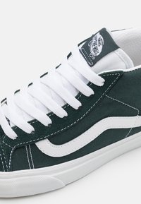 Baskets Vans vert foncé avec bande latérale blanche, lacets blancs et logo Vans "Off The Wall" sur la languette sur fond blanc.