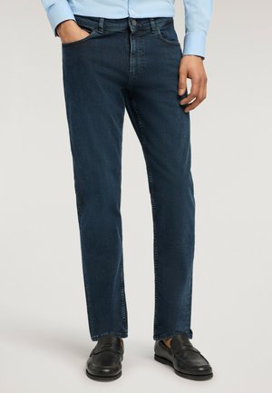 Mies, jolla on tummansiniset suorat farkut, vaaleansininen paita ja mustat nahkaiset loaferit, seisoo yhdellä kädellä taskussa.