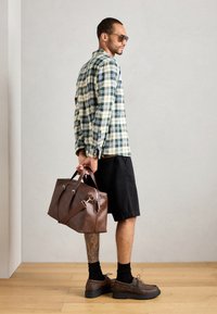 Plaid button-up shirt in green and cream, black shorts, brown leather bag with handles and strap, brown shoes, and sunglasses.