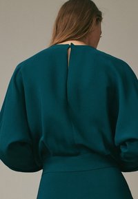 Donna con lunghi capelli castani che indossa un abito verde acqua con maniche voluminose e una piccola apertura a chiave sulla schiena vicino al colletto.