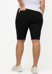 Pantaloni corti in denim nero con un design aderente, lunghezza a metà coscia, tasche posteriori e lievi sfilacciature all'orlo, abbinati a sneakers bianche.