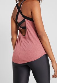 Débardeur à dos croisé en tissu rouge rosé. Présente un design ajouré, des bretelles noires contrastantes et une texture douce. Associé à des leggings noirs.