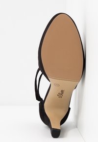 Chaussure à talon haut en daim noir avec un dos ouvert, une fine sangle autour de la cheville et un bout pointu. La semelle est couleur tan avec une finition texturée et un marquage.