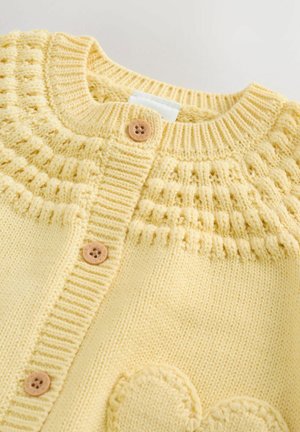 Vaaleankeltainen neulottu neuletakki, jossa on ribbireuna, teksturoitu yläpää, kolme puista nappia ja pyöreä, kohotettu sydänkoriste.