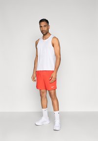 Άσπρο φανελάκι χωρίς μανίκια, κόκκινα αθλητικά σορτς με λευκό λογότυπο Nike, λευκά αθλητικά παπούτσια και λευκές κάλτσες, σε απλό φόντο.