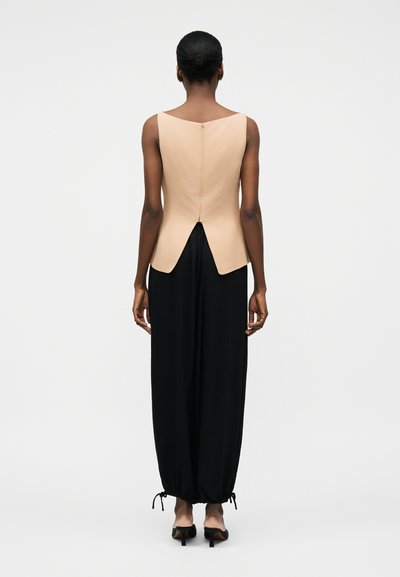 Žena stoji okrenuta leđima prema naprijed, nosi bezrukavu bež majicu sa zatvaračem na leđima i crne široke hlače s vezanim rubovima te crne štikle.