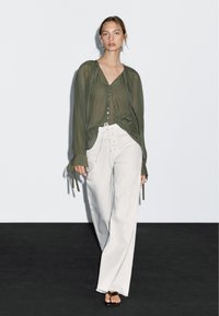 Blusa verde oliva trasparente con chiusura a bottoni e maniche legate, abbinata a pantaloni bianchi a gamba larga con laccio frontale e orli sfilacciati.