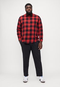 Camisa de franela a cuadros rojos y negros, de manga larga y con cierre de botones, combinada con pantalones negros y zapatillas blancas.