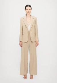 Beige op maat gemaakte suit bestaande uit een enkelzijdige blazer en brede broekspijpen. Het jasje heeft klepzakken en gelapte revers.