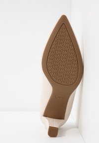 Chaussure à talon beige à bout pointu montrant une semelle marron avec un motif répété de logo "MK" et un petit talon carré.