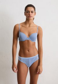 Mujer vistiendo un sujetador acolchado azul claro y ropa interior de encaje a juego, de pie contra una pared blanca lisa con una expresión neutral.