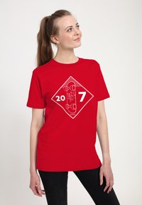Ung kvinde iført en rød t-shirt med et hvidt skateboard-tryk og tallene 20 og 7, kigger opad til højre.