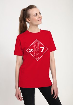 Ung kvinde iført en rød t-shirt med et hvidt skateboard-tryk og tallene 20 og 7, kigger opad til højre.