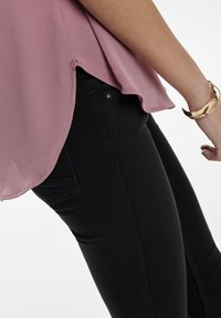 Blusa texturizzata rosa pallido con orlo curvo, abbinata a jeans in denim neri con tasca posteriore e dettagli dei bottoni visibili.