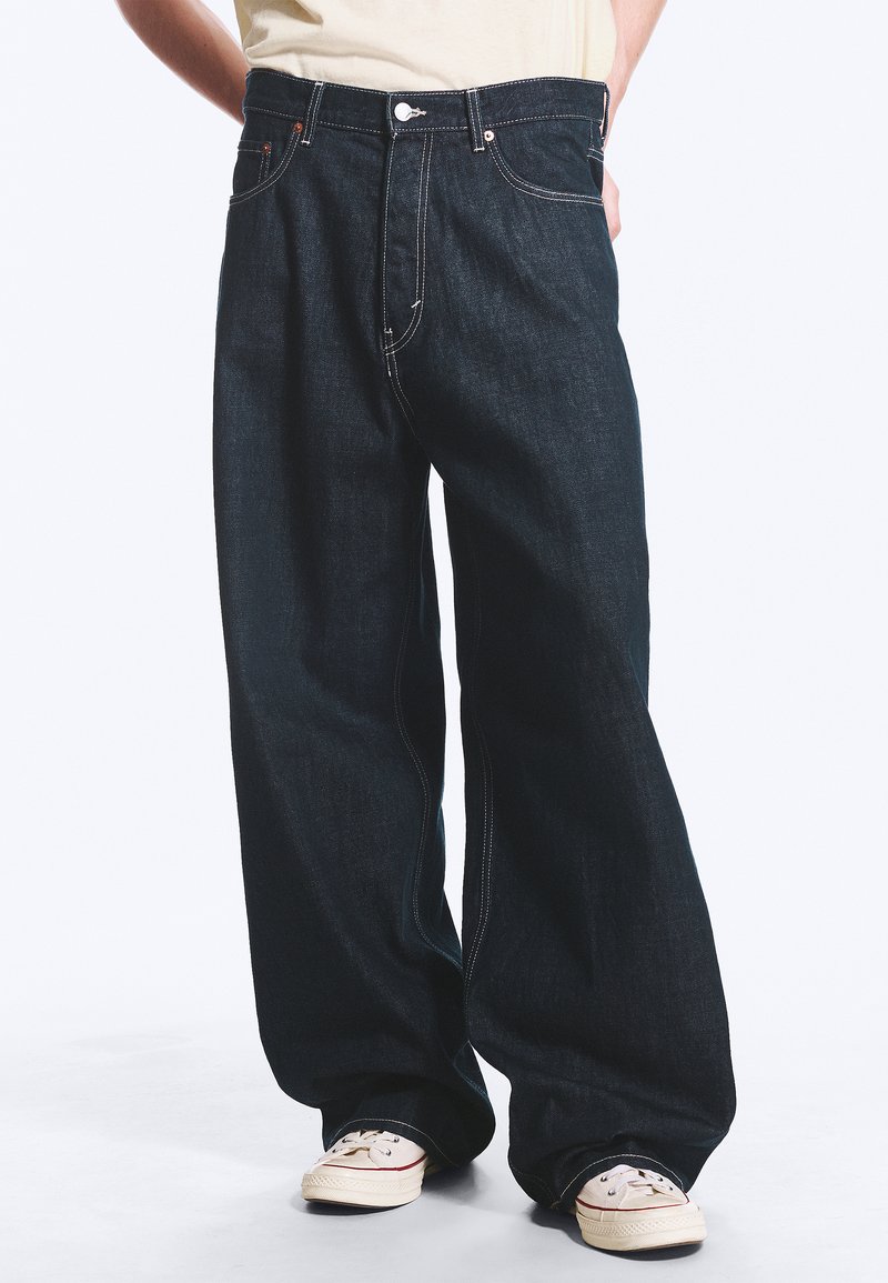 Persona che indossa jeans blu scuro larghi e sneakers bianchi, mani nelle tasche, in piedi davanti a uno sfondo chiaro e semplice.
