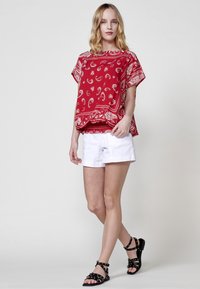 Top corto rosso a maniche corte con motivo paisley bianco, abbinato a pantaloni corti bianchi e sandali neri con cinturini. Tessuto morbido, taglio rilassato.