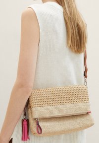 Tkaná slámová crossbody taška s plochým designem. Má růžovou třásničku a fialové akcenty, zipovou přihrádku a hnědý popruh.