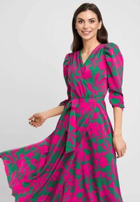 Sukienka w kwiaty z zielonym tłem i wyrazistymi różowymi wzorami, z bufiastymi rękawami i związanym w pasie, co podkreśla sylwetkę.