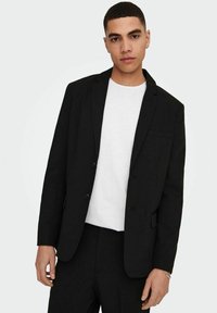 Blazer nero con design a un bottone, rever a punta, due bottoni frontali e taschino sul petto. Indossato su una maglietta bianca.