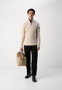 Jersey beige con cuello alto y textura acanalada, pantalones negros, zapatos negros y una bolsa de lona marrón claro con acentos marrones.