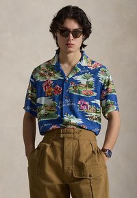 Jeune homme portant des lunettes de soleil et une chemise à imprimé tropical bleu avec un pantalon marron taille haute et une montre-bracelet, debout devant un fond beige.