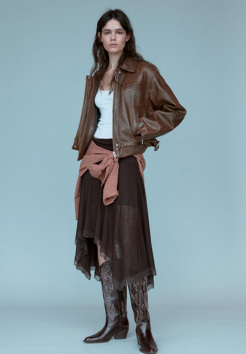 Jeune femme portant une veste en cuir marron, un débardeur blanc, une jupe asymétrique marron, une chemise nouée et des bottes de cowboy brodées foncées.