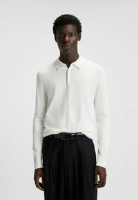 Polo shirt bianco a maniche lunghe con colletto, realizzato in tessuto liscio, abbinato a pantaloni neri e una cintura nera elegante.