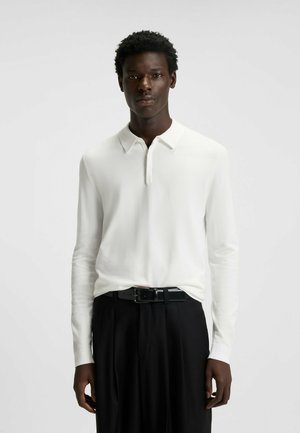 Polo à manches longues blanc avec un col, en tissu lisse, associé à un pantalon noir et une ceinture noire élégante.