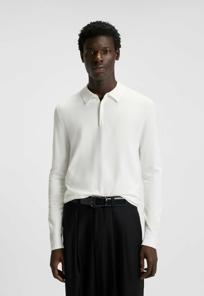 Polo shirt bianco a maniche lunghe con colletto, realizzato in tessuto liscio, abbinato a pantaloni neri e una cintura nera elegante.