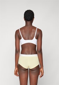 Bralette blanche avec des bretelles ajustables associée à un short hipster jaune avec une taille blanche portant l'inscription "PIECES" en lettres jaunes.