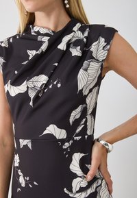 Rochie neagră cu model floral alb, cu o decolteu drapat, mâneci scurte și un material neted, texturat. Brățară argintie accentuată pe braț.