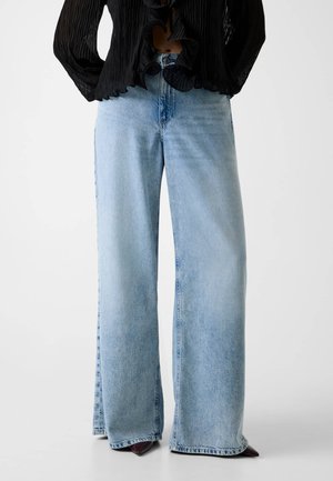 Persona che indossa jeans azzurri a gamba larga e una camicetta nera testurizzata con bordi arricciati, in piedi su uno sfondo bianco.