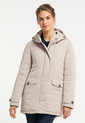 Donna che indossa un cappotto invernale beige chiaro con cappuccio e tasche con bottoni sopra un maglione scuro, in piedi davanti a uno sfondo bianco uniforme.