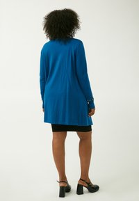 Tunic blu con maniche lunghe e taglio dritto, che arriva a metà coscia. Presenta polsini testurizzati con dettagli decorativi. Gonna nera e tacchi spessi.