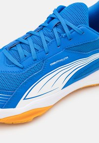 Blå atletiske sko med mesh-materiale, hvide detaljer, stabilitetsbranding og en struktureret orange gummi sål.