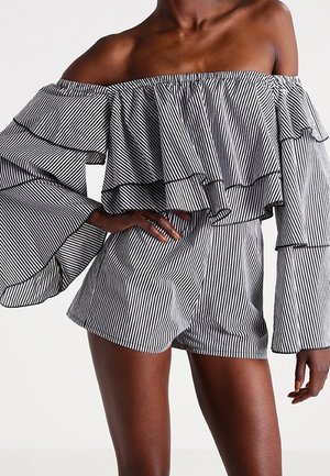 Vrouw draagt een zwart-wit gestreepte off-shoulder romper met een gerimpelde top en wijde lange mouwen, staand met één hand op de heup.