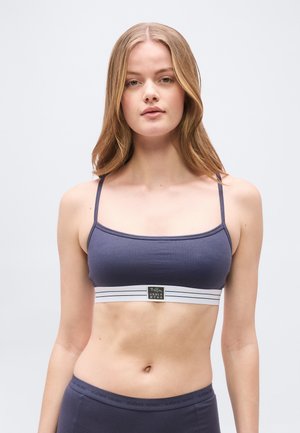 Young woman with light brown hair wearing a dark blue sports bra and matching high-waisted bottoms against a plain gray background.