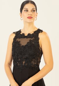 Schwarzes Kleid mit ärmellos Design, verziert mit detaillierter schwarzer Spitze und Perlen auf einem transparenten Brustbereich. Glatter Stoff unterhalb der Taille.