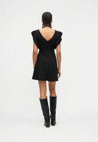 Vrouw met gevlochten haar, gekleed in een korte zwarte jurk met ruches aan de mouwen en zwarte laarzen tot aan de knie, staand met haar rug naar voren op een witte achtergrond.