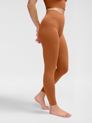 Kvinde iført brune, ribstrikkede leggings med høj talje og matchende kort top, stående barfodet med hænderne bag ryggen mod hvid baggrund.